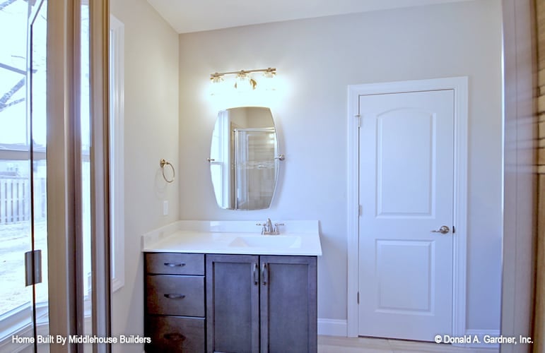 A closer look at one of the primary bathroom's vanity with dark wood cabinets, a porcelain sink, and a frameless mirror well-lit by a glass sconce.