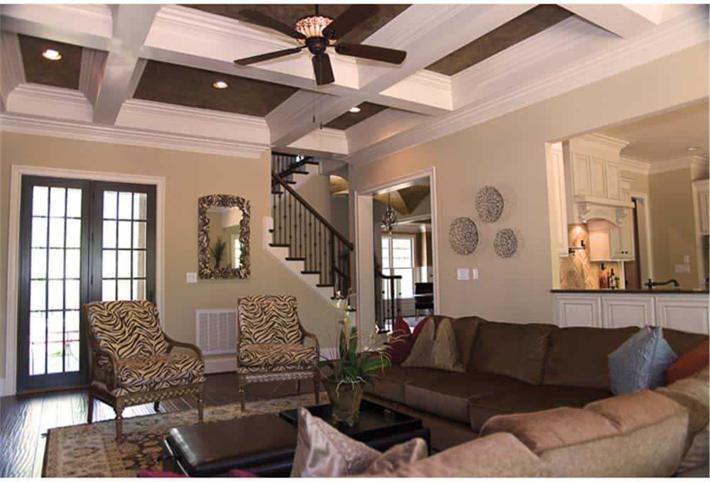 Great room with patterned armchairs, an L-shaped sectional, and a ceiling fan mounted on the coffered ceiling.