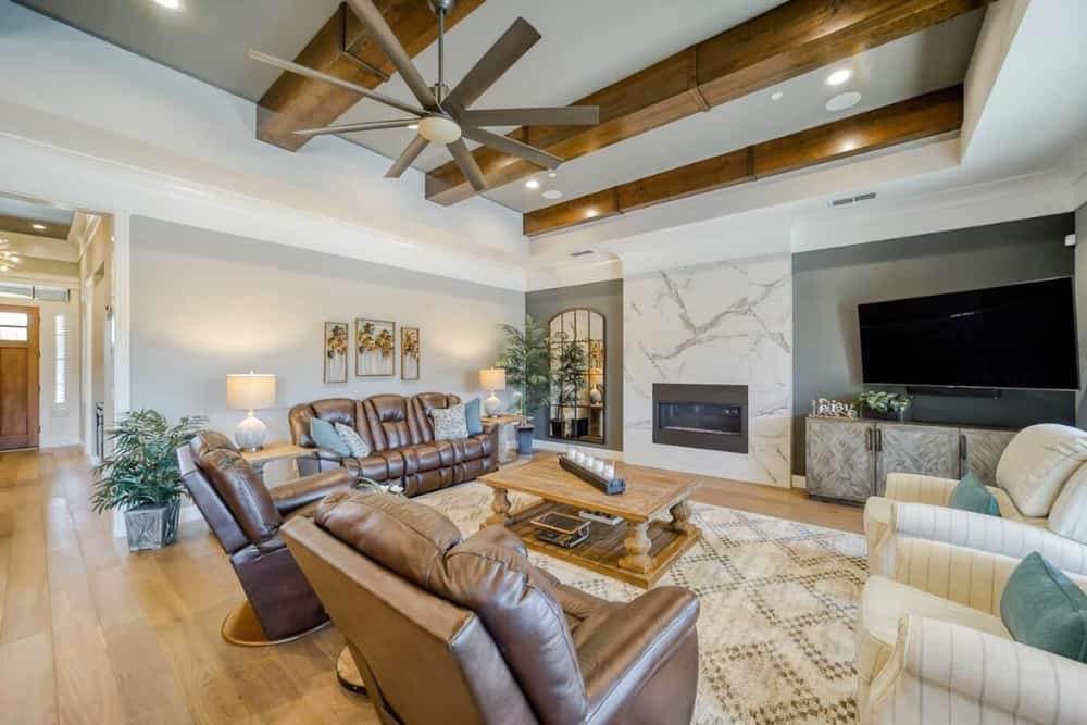 Great room with leather and fabric seats, a wooden coffee table, a marble fireplace, a TV, and an industrial fan hanging from the beamed ceiling.