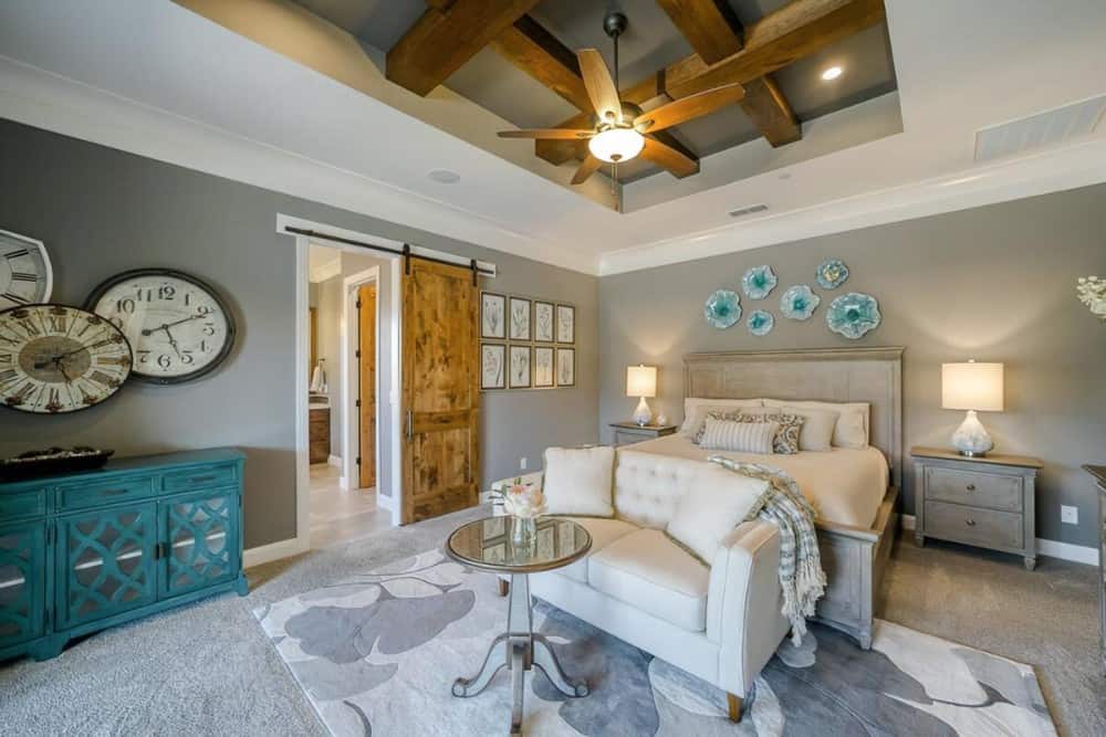 The primary bedroom features a tray-beamed ceiling, carpet flooring, and gray walls adorned with clocks and floral wall arts.