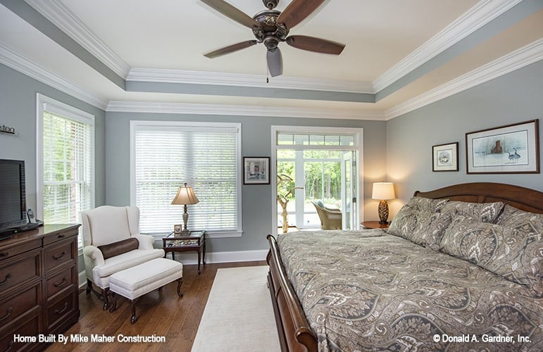 The primary bedroom features a tray ceiling, natural wood furnishings, a white wingback lounge chair, and private access to a covered porch.
