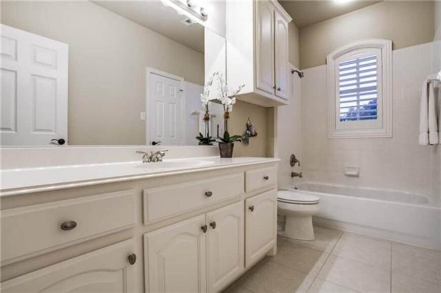 This bathroom is equipped with white vanity, a toilet, and a tub and shower combo brightened by a louvered window.