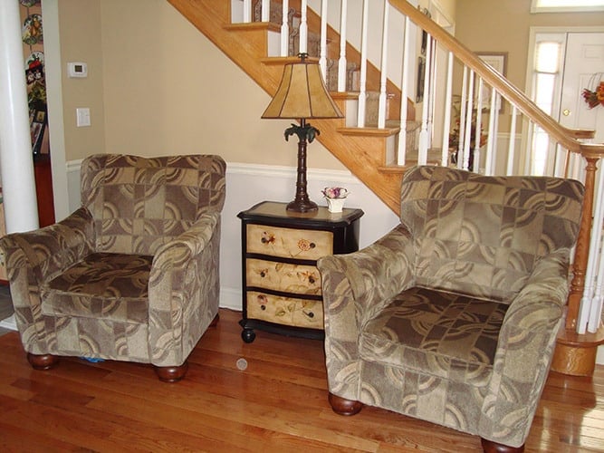 The foyer includes patterned armchairs and a side table placed against the staircase.
