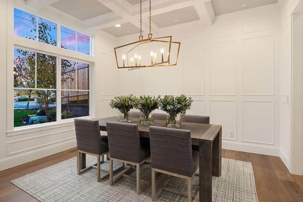 Formal dining room with full height wainscoting, coffered ceiling, and a rectangular dining set well-lit by a brass chandelier.