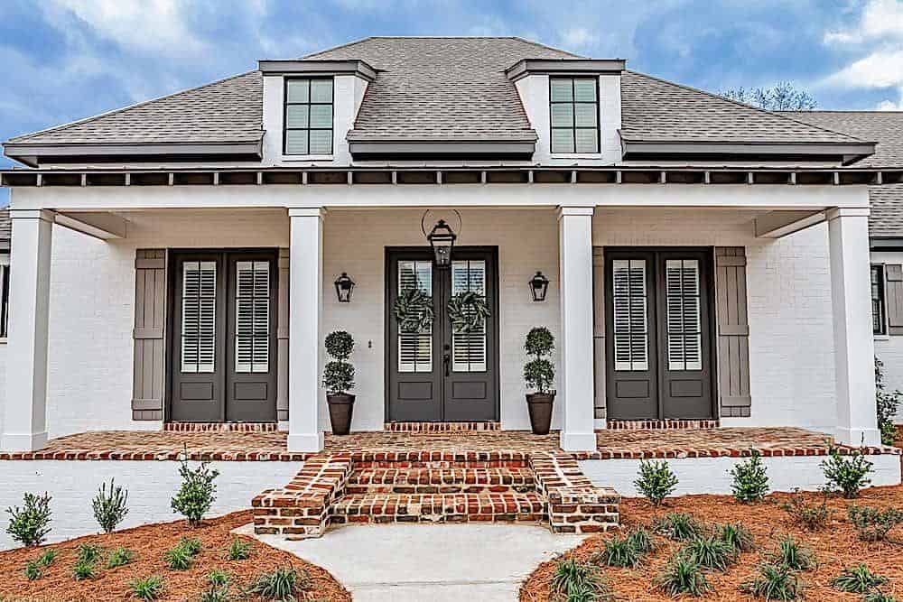 A closer look at the covered front porch with three french doors, white columns, brick flooring, and a stoop.