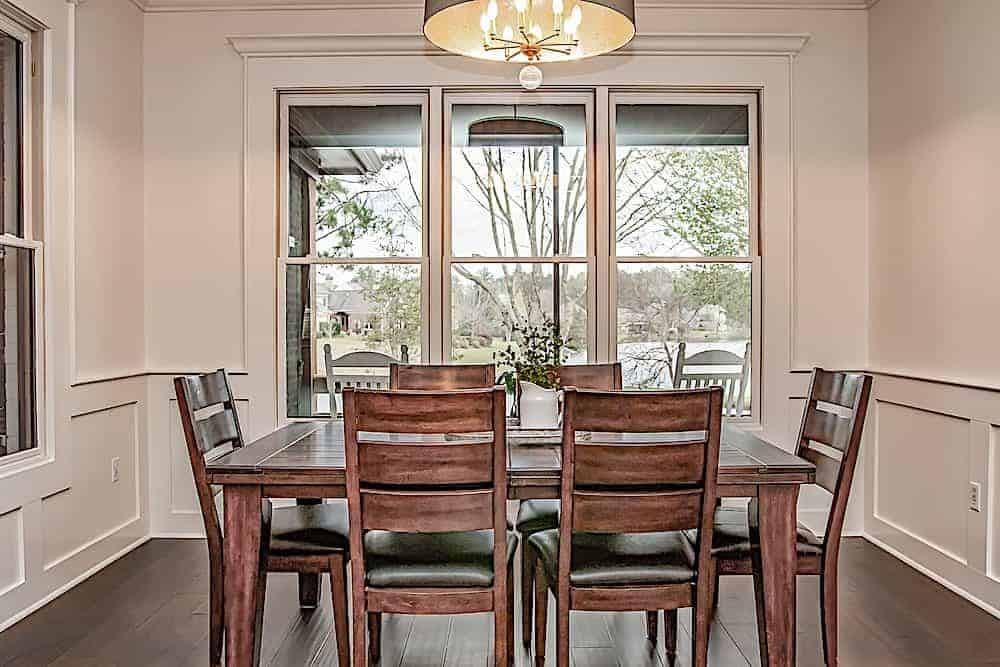 Dining area with white wainscoted walls, large windows, and a wooden dining set well-lit by a drum pendant.