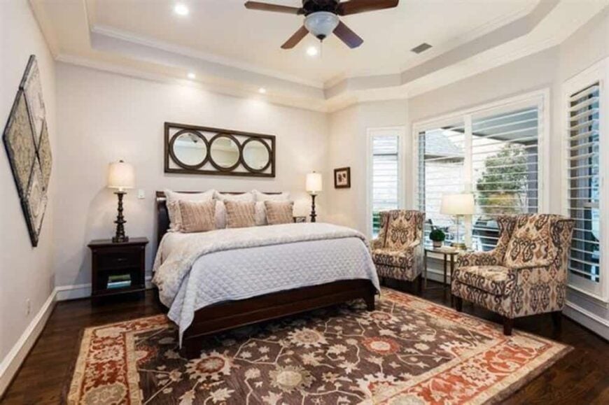 The primary bedroom features a tray ceiling and a sitting area by the bay window furnished with patterned wingback chairs.