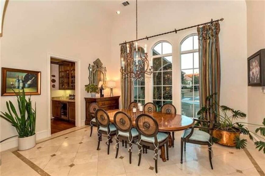 The formal dining room has an oval dining set, fresh potted plants, and tall arched windows dressed in striped drapes.