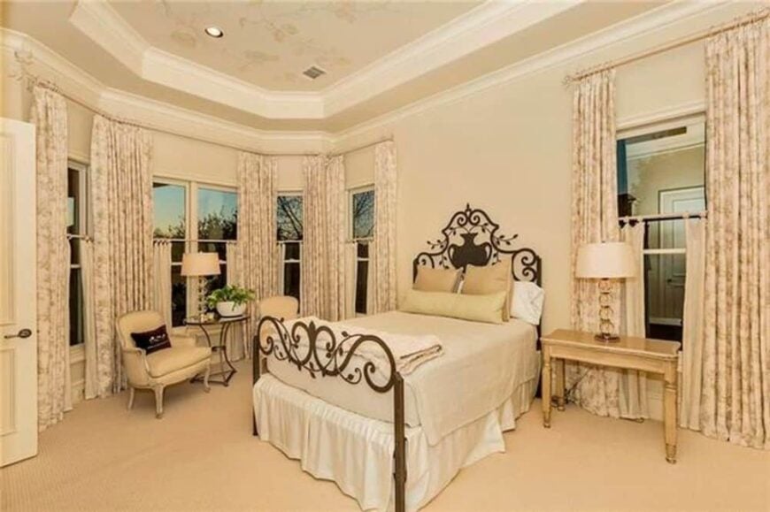 This bedroom features an elegant tray ceiling, surrounding windows, and an ornate bed complemented with a wooden nightstand and cushioned armchairs.