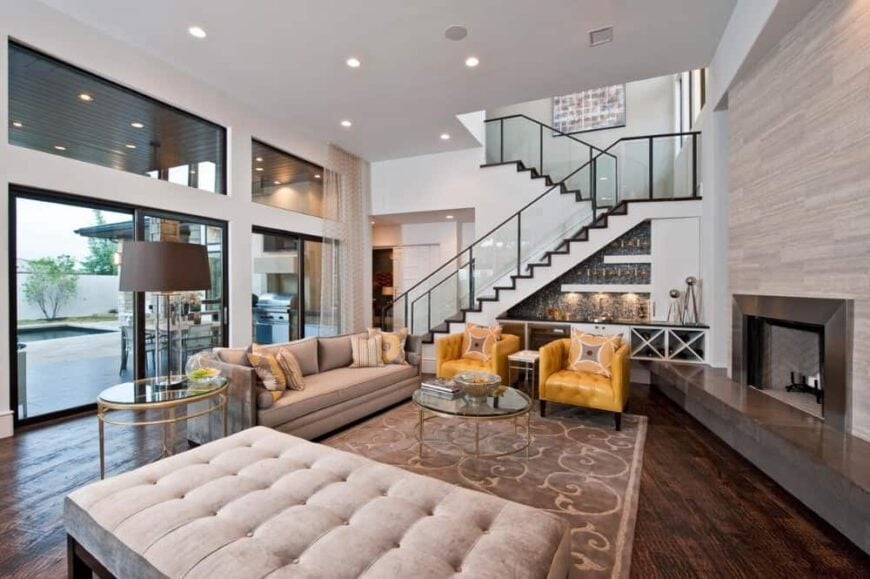 The great room is filled with a gray sofa, a large ottoman, leather tufted chairs, a modern fireplace. and a wet bar.