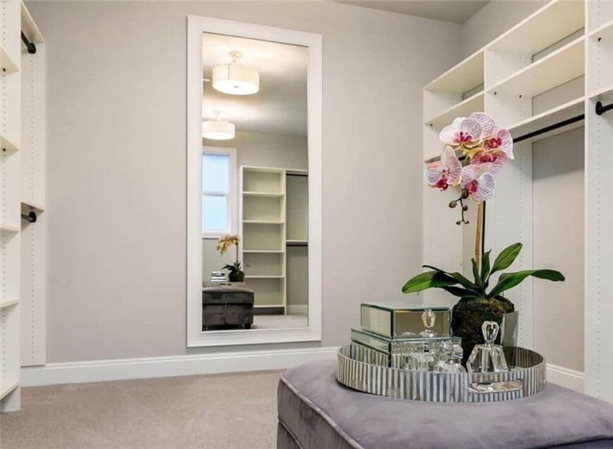 The primary walk-in closet is filled with white built-in shelves, a full-length mirror, and a velvet ottoman topped with a round tray.