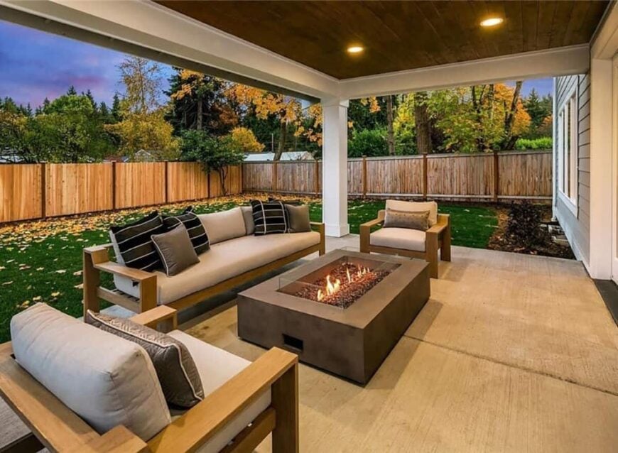 Covered porch with cushioned seats and a fire pit table. It transitions to a lush lawn bordered by a wooden fence.