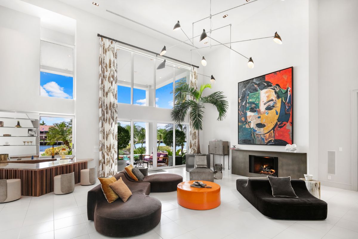 Two story living area with full lenght glass wall, cushioned sofas, and concrete fireplace connected to bar area.