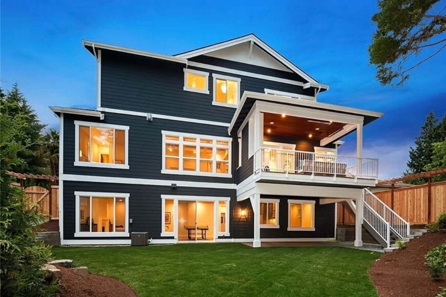 Rear exterior view of the house showcasing its dark siding, covered porches, and plenty of windows highlighted by ambient lighting.