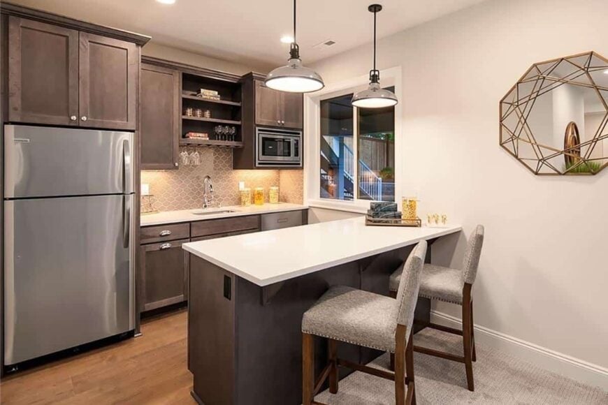 The wet bar is equipped with slate appliances, wooden cabinets, and a peninsula paired with gray upholstered chairs.