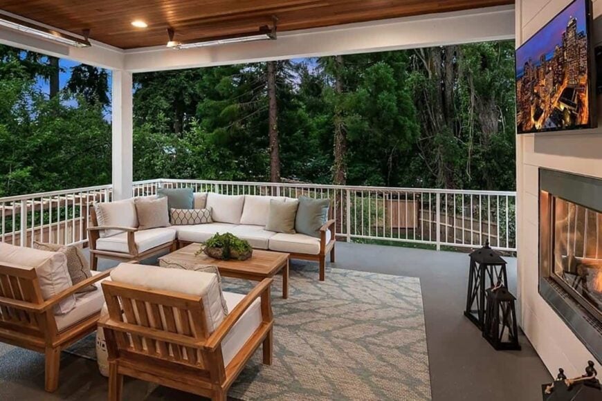 The covered deck is filled with cushioned seats, a wooden coffee table, and a fireplace topped with a wall-mounted TV.