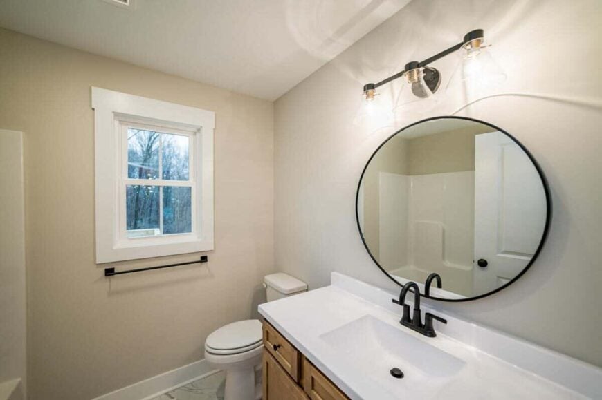 This bathroom offers a toilet, a sink vanity, and a tub and shower combo reflected in the round mirror.