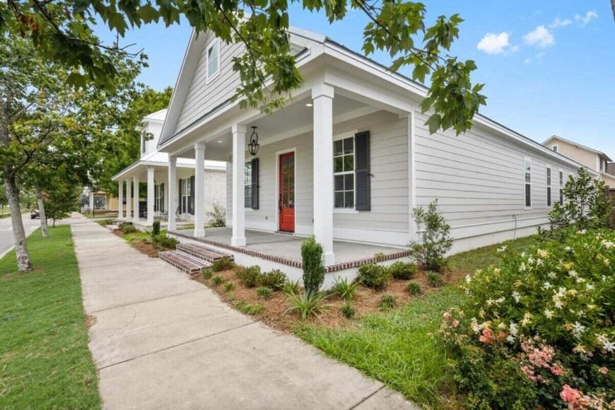 Angled front rendering showcasing a horizontal lap siding, shuttered windows, a red front door, and a covered porch lined with white pillars.