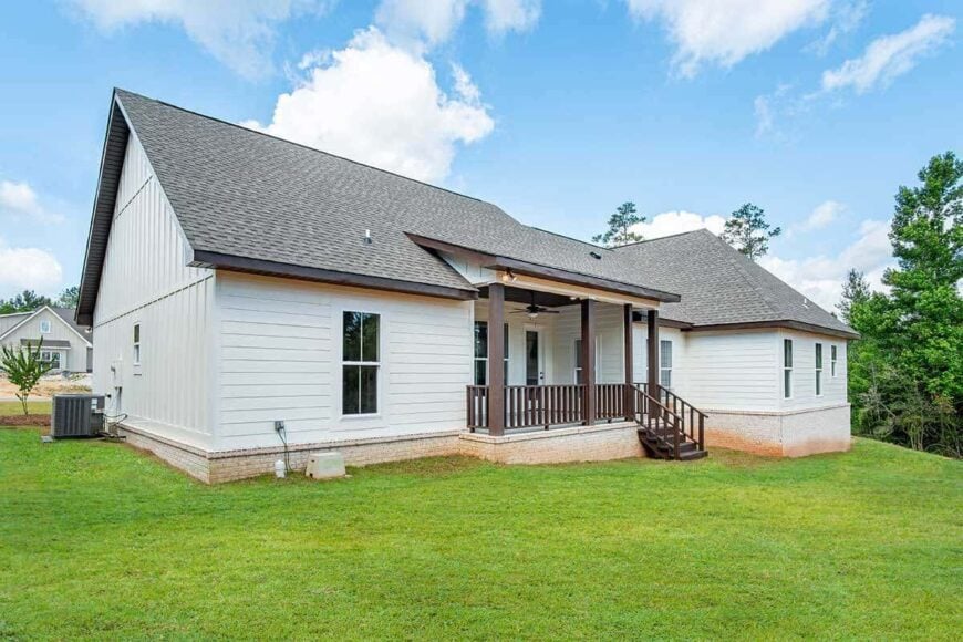 View of the house from the backyard showcasing its white siding, brick skirting, and a covered porch bordered by wooden columns and railings.