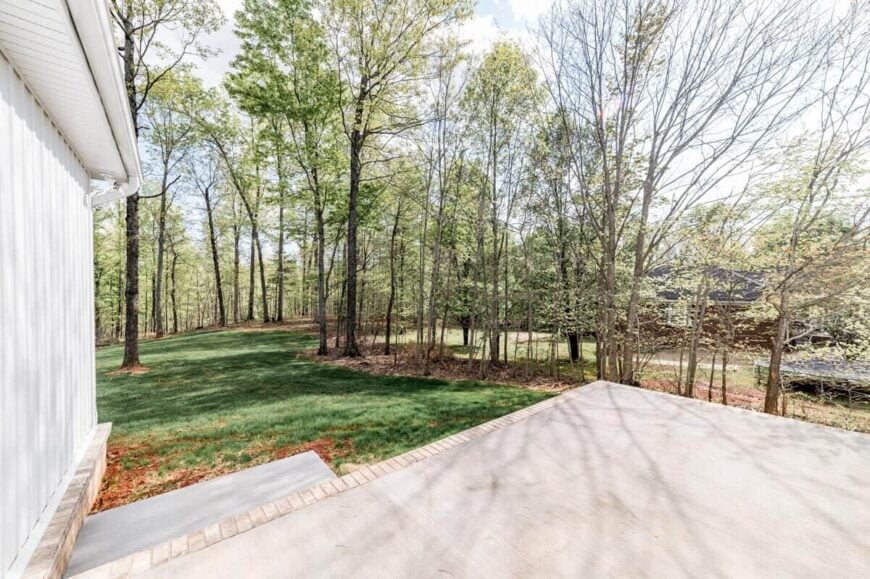 An open patio at the back with a concrete floor bordered by bricks. It transitions to a lush lawn with towering trees.