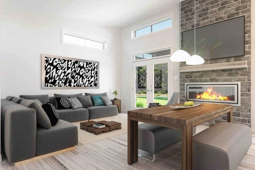 Combined living and dining room with gray seats, transom and clerestory windows, a wooden dining set, and a large TV fixed above the fireplace.