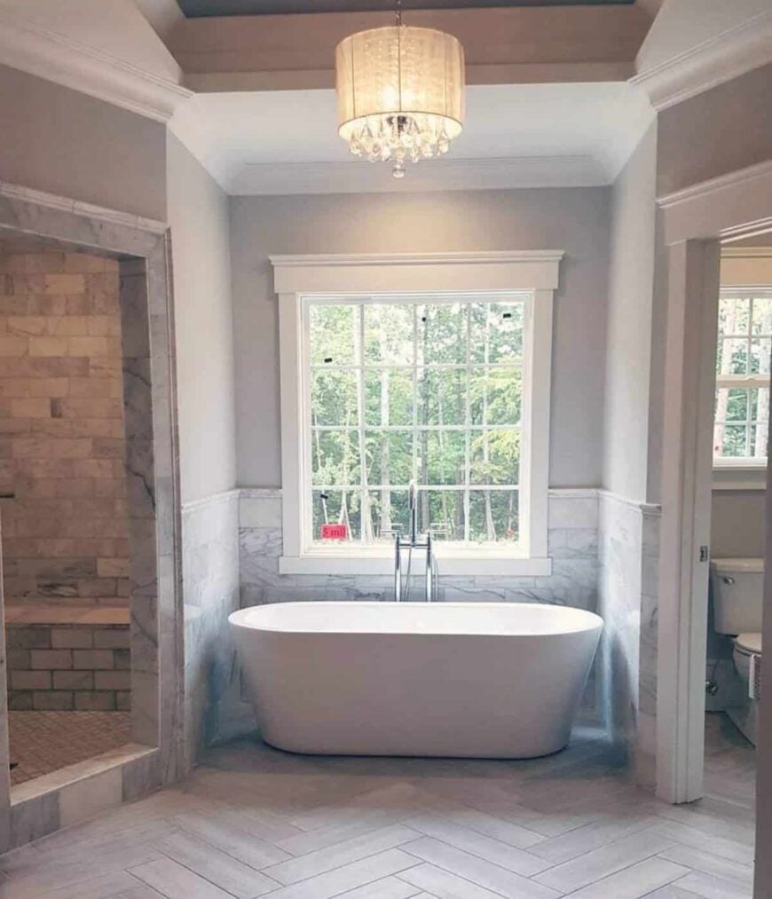 The primary bathroom features a freestanding tub well-lit by a framed window and an elegant drum chandelier.