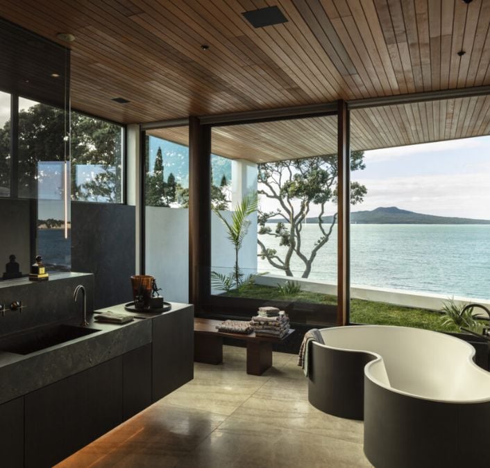 An interior shot of a modern bathroom design, featuring a freestanding bathtub as the centerpiece and showcasing the stunning oceanic view through the sliding glass walls.