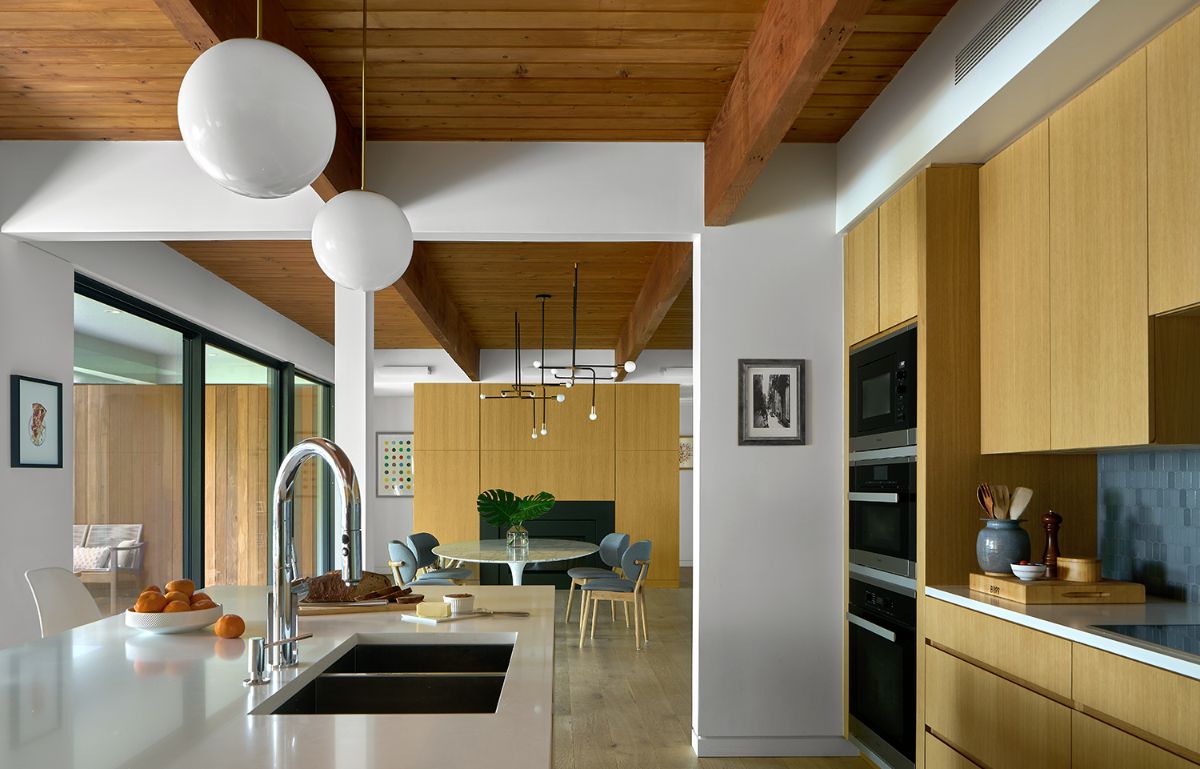 After-renovation kitchen and dining room with oak wood cabinets and beamed ceiling.