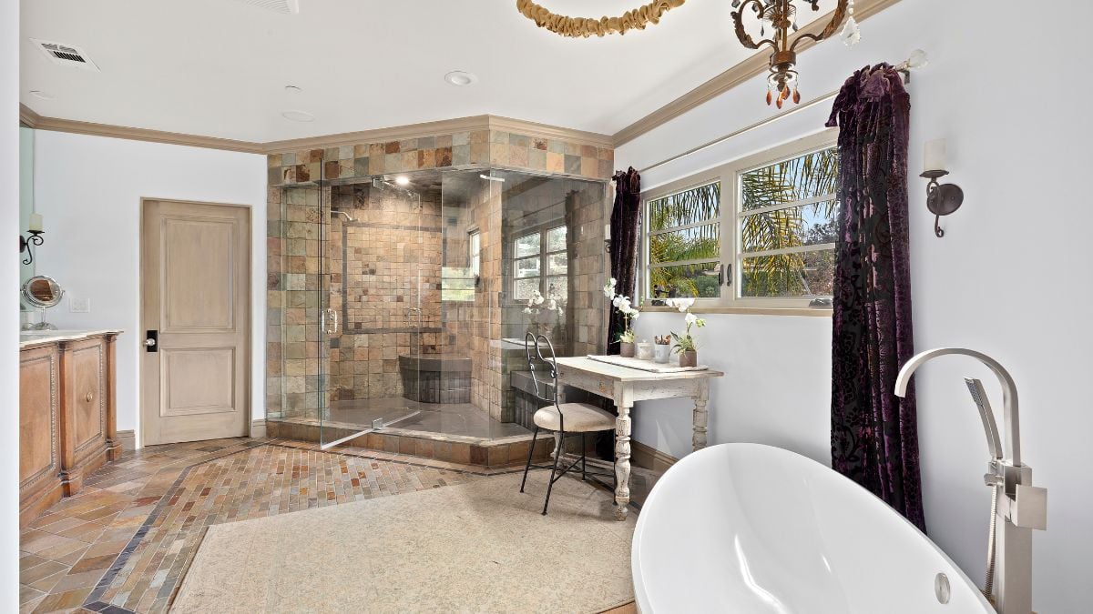 This bathroom features a separate shower area, a white bathtub, and a small wooden table and chair close to the window.