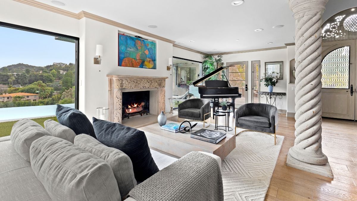 The living room features one gray sofa, two dark gray chairs, a carpet underneath, a pillar on one side, a fireplace on the other, and a piano at the back.