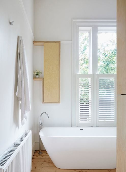 This is an interior photograph of a bathroom showcasing a white, clean and minimalist design with a bathtub as the central feature.