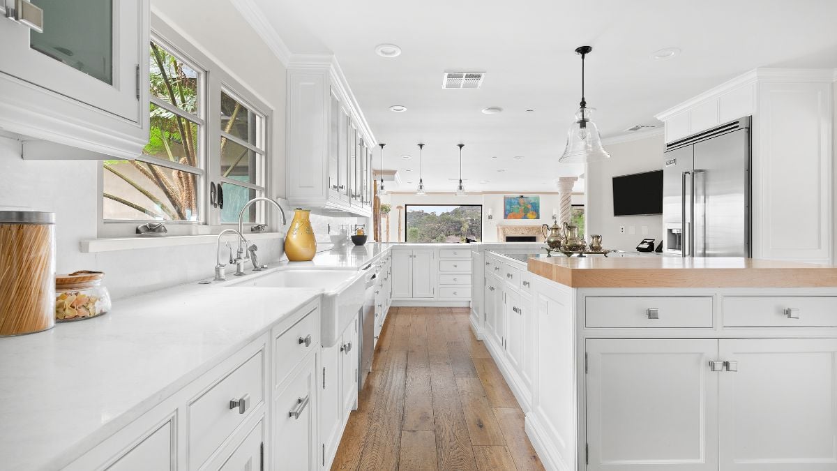 This kitchen area is all white, features wooden floors, all the necessary appliances, and views out the windows.