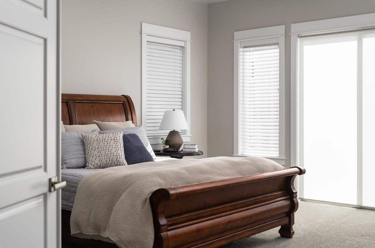 The primary bedroom has carpeted floor, a wooden bed, and tall windows covered in rolling blinds and shades.