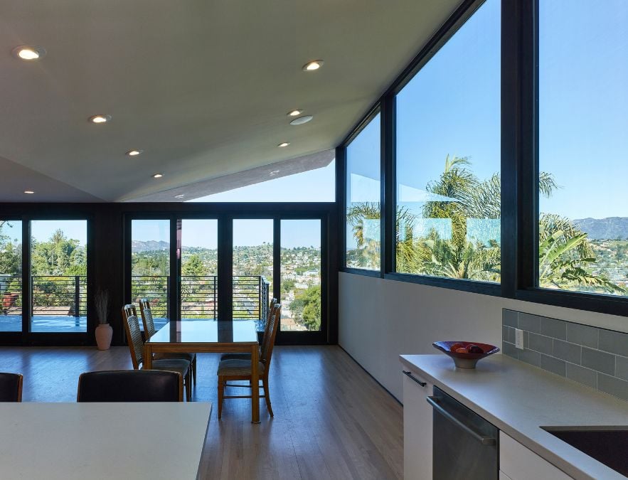 This photograph captures a kitchen and dining room area, surrounded by glass doors and windows that offer a scenic view of the surrounding outside environment.
