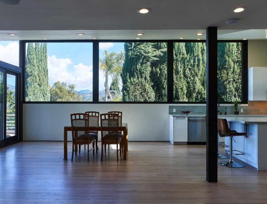 A dining area consisting of a table and chairs is situated in front of expansive glass windows, affording a captivating view of the outdoors, with a focus on the impressive height of the surrounding trees.