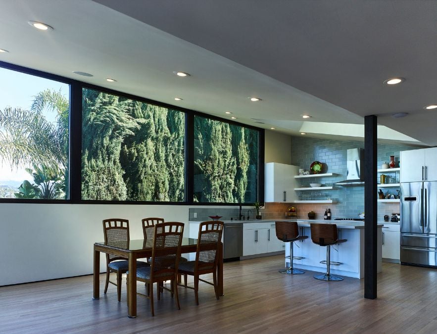 This residence features a kitchen and dining area that is strategically situated in front of expansive glass windows which offers a view of the surrounding environment includes towering trees.