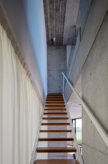 A staircase located near the wall leads to the upper level, which features a minimalist white design complemented by raw concrete.