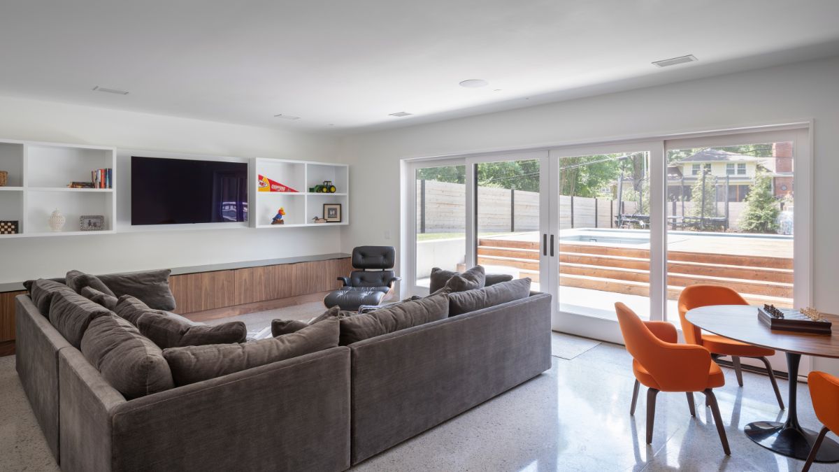 The living area, which has cushioned sofas, open shelving, a large TV, and a sliding door leading to the backyard with a swimming pool.