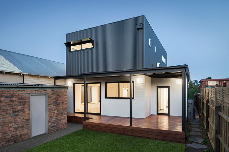 The house's back facade, with white and dark cladding, a wooden deck, and a small lawn.