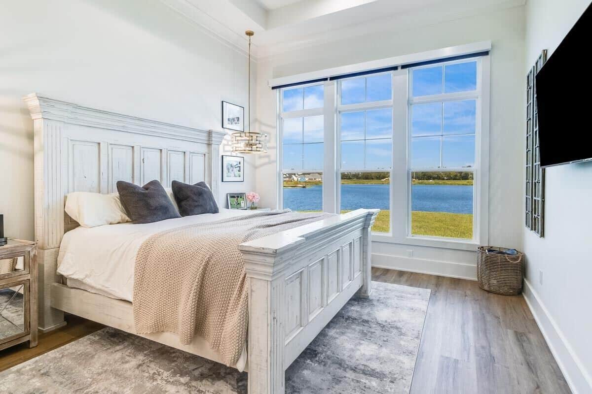 Primary bedroom with a distressed white bed and a trio of windows overlooking the surrounding landscape.