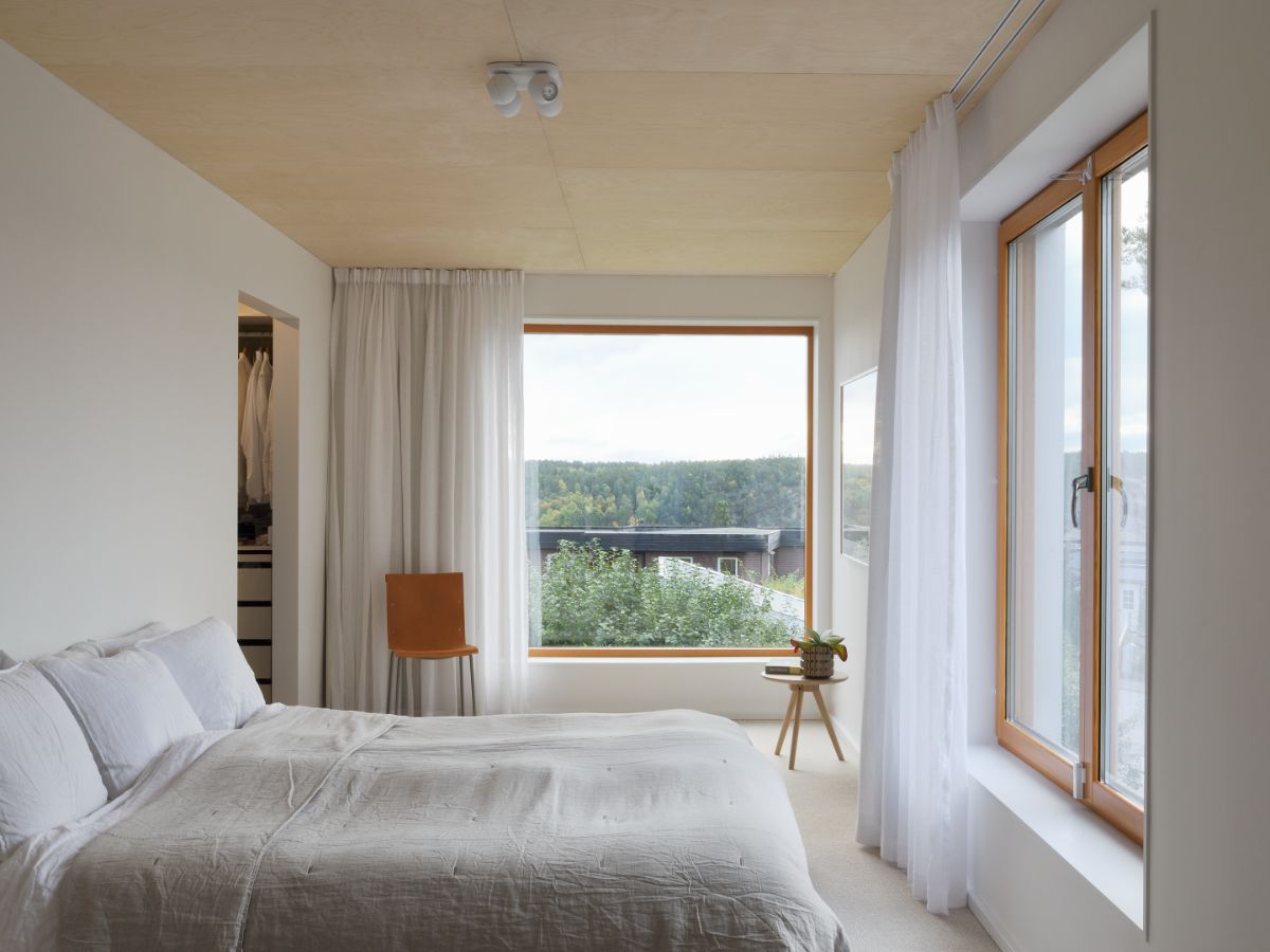 A bedroom with large windows draped in white sheer curtains giving a view of the neighboring houses.