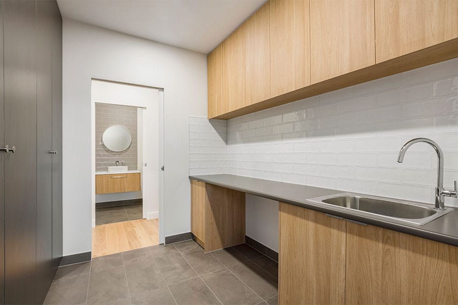 The laundry room, which has wooden cabinetry, a metal sink with a white brick backdrop, and a floor to ceiling closed organizer.