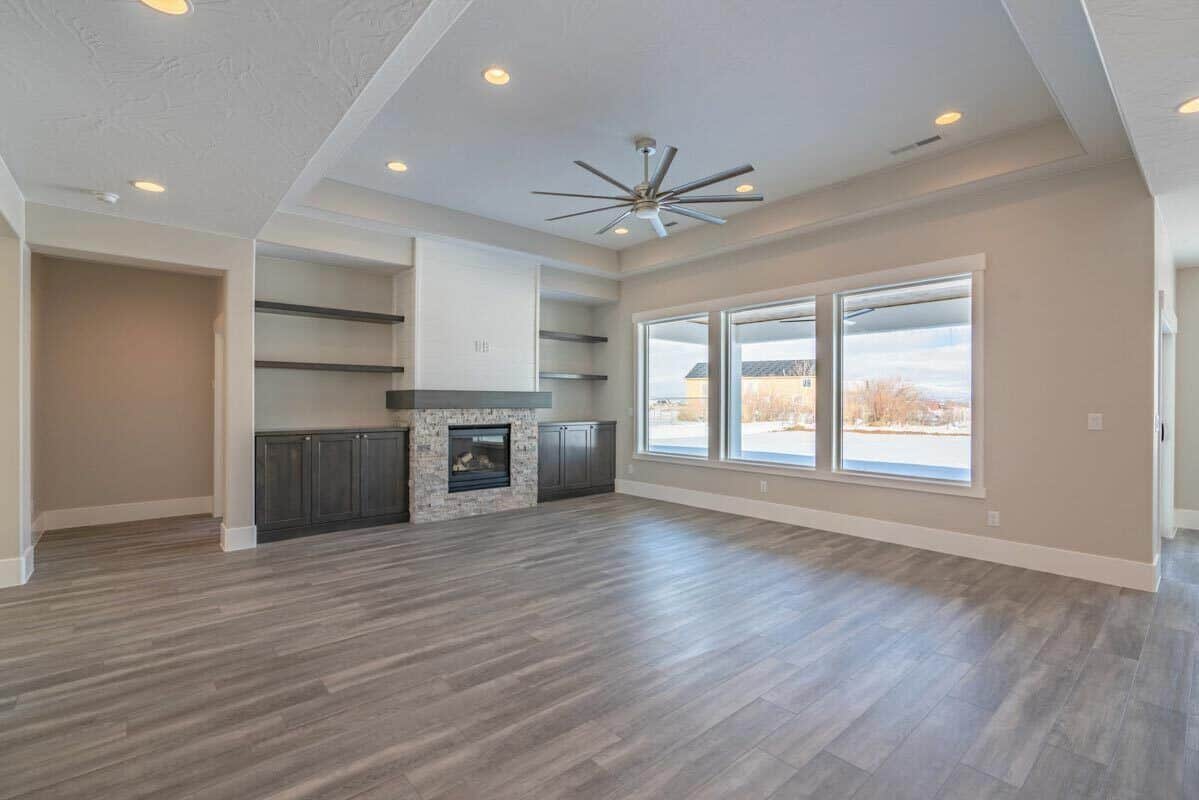 The great room has a tray ceiling, a trio of windows, and a stone fireplace flanked by floating shelves and base cabinets.