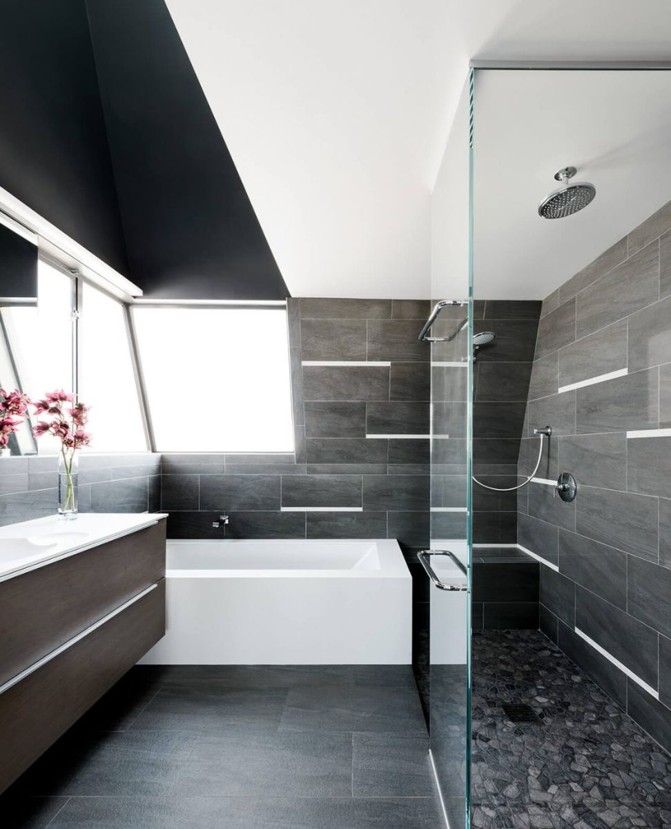 The use of grey and white in this bathroom creates a modern look. The white alcove bathtub contrasts with the black wall, and the walk-in shower is surrounded by glass with pebble floors. The ceilling is dark and white in color.