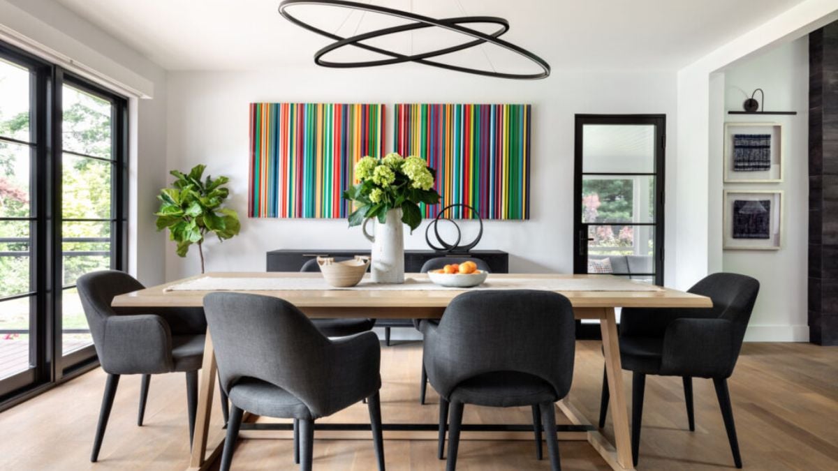 The dining room is decorated artistically and features black chairs, a black modern chandelier, a flower arrangement on top of the hardwood table, and a beautiful view of the artwork on the wall in the space directly in front of it.
