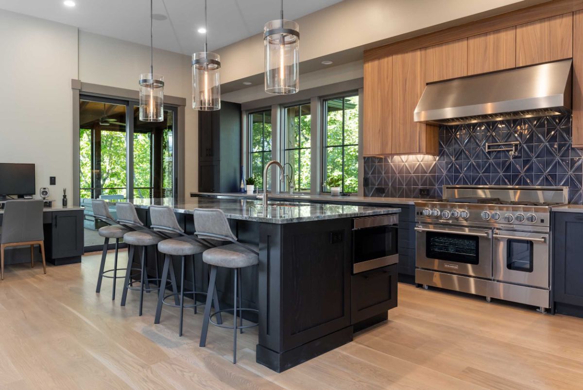 The kitchen features modern components such as a marble island, high-end appliances, and modern pendant lighting. The dark cabinetry stands out against the natural wood floors.