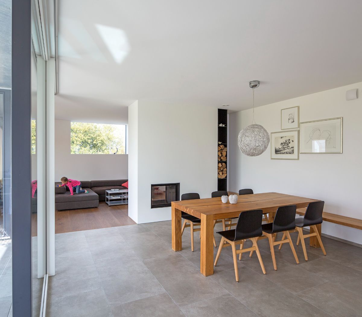 A double-sided fireplace separates the dining and living areas. The dining room is distinguished by black furniture fronts and concrete-colored tiles, whereas the living room is made of rustic oak wood with built-in furniture and floors.