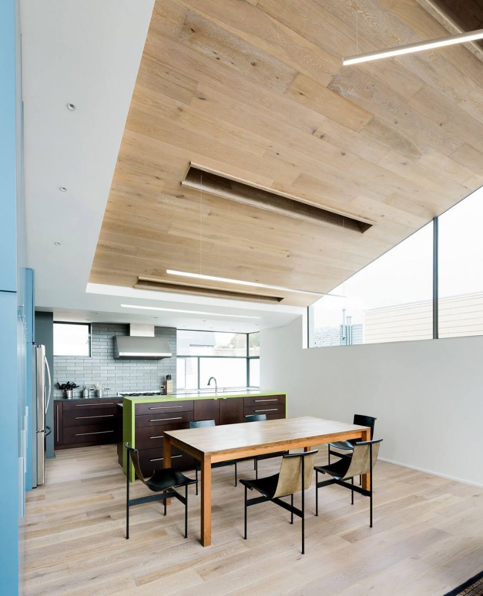 The dining and kitchen area in the sky house unit has a light blue wall built-in cabinets, and the mix of green and dark brown in the island contrasts nicely. Natural wood flooring that extends all the way to the ceiling complements the wooden dining area.