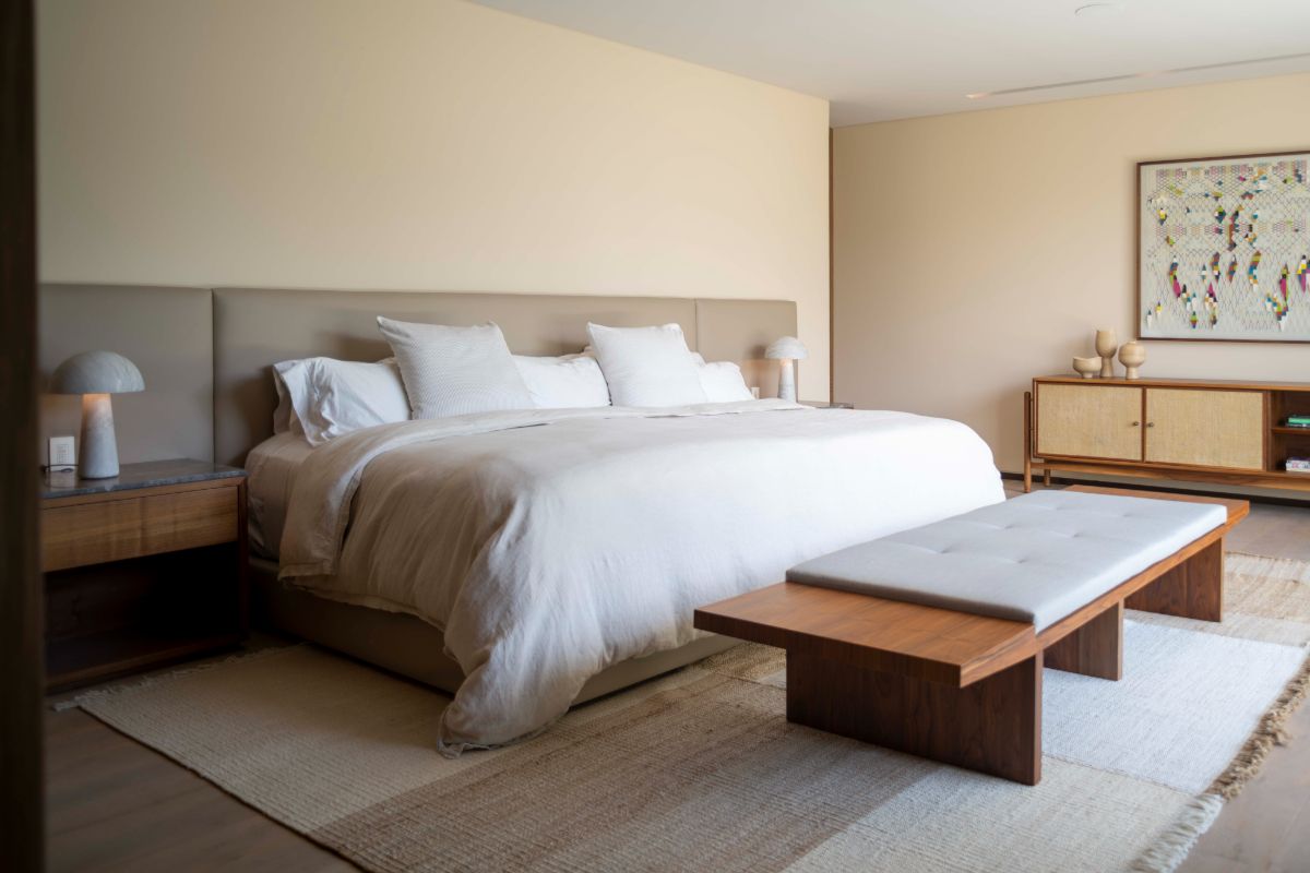The bedroom has a rug, a wooden bedroom seat, and wooden cupboards. The flooring is made of light wood, and the walls are painted in a cream tone.