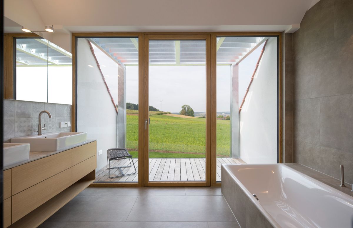 A light-filled bathroom with a deep soaking tub and a counter-topped dual sink on floating cabinetry with a full-width mirror. The flooring is made of concrete-colored tiles that extend all the way to the wall. It features a floor-to-ceiling glass window with a door leading to a semi-covered deck.